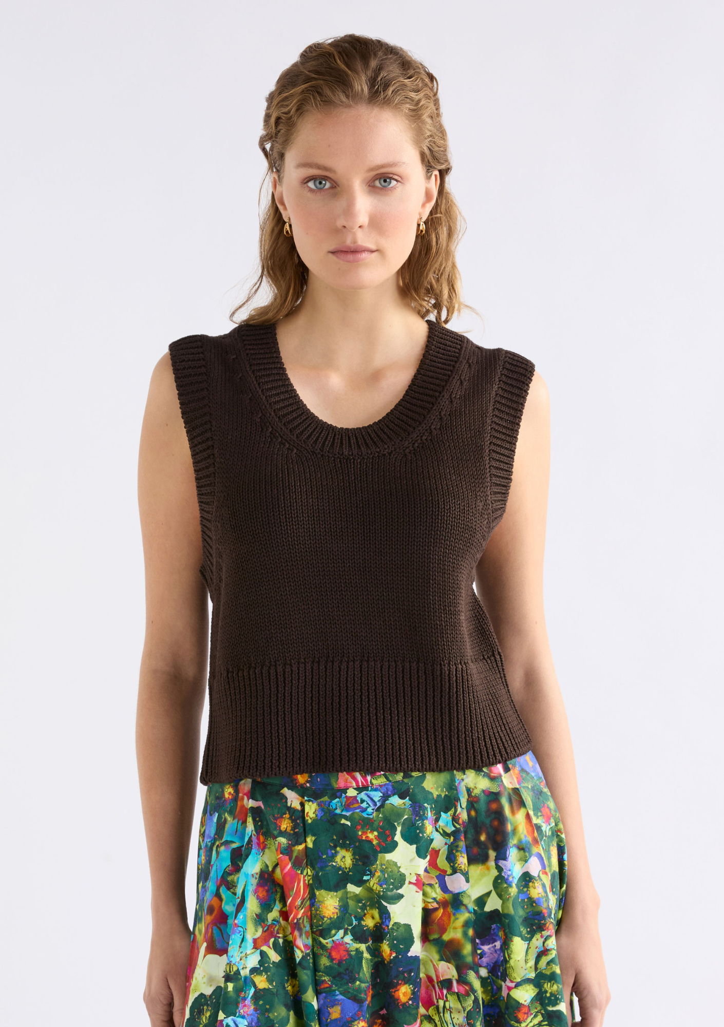 Woman wearing a brown sleeveless knitted top with a colorful skirt on a white background